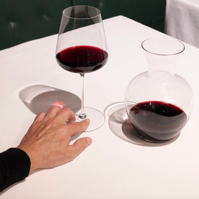 Hand holding glass of red wine with a carafe of red wine on a white table beside him