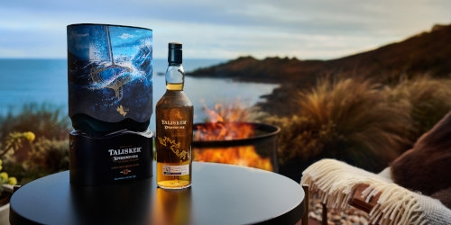 Bottle and box on table with the sea, sunset, and fire pit behind